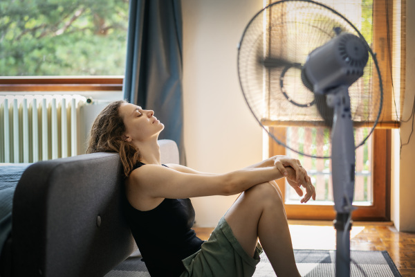 Junge Frau, die unter Hitze leidet, sitzt zu Hause auf dem Boden vor einem Ventilator. © PantherMedia/OlezzoSimona