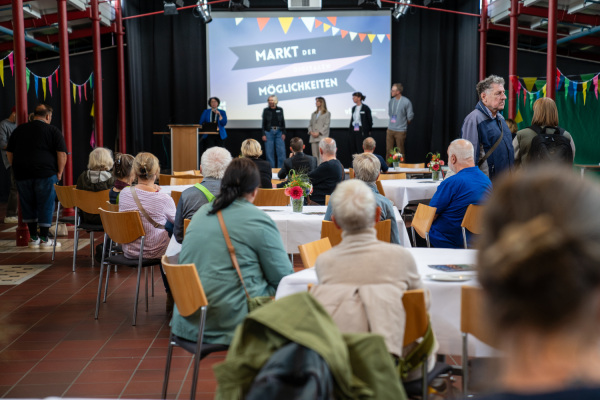 Menschen sitzen beim Markt der digitalen Möglichkeiten an den Tischen und hören den Organisator*innen auf der Bühne zu.