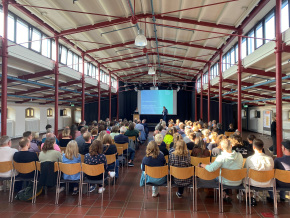 Plenum mit 140 Menschen, auf der Bühne zwei Männer, auf dem Bildschirm steht 